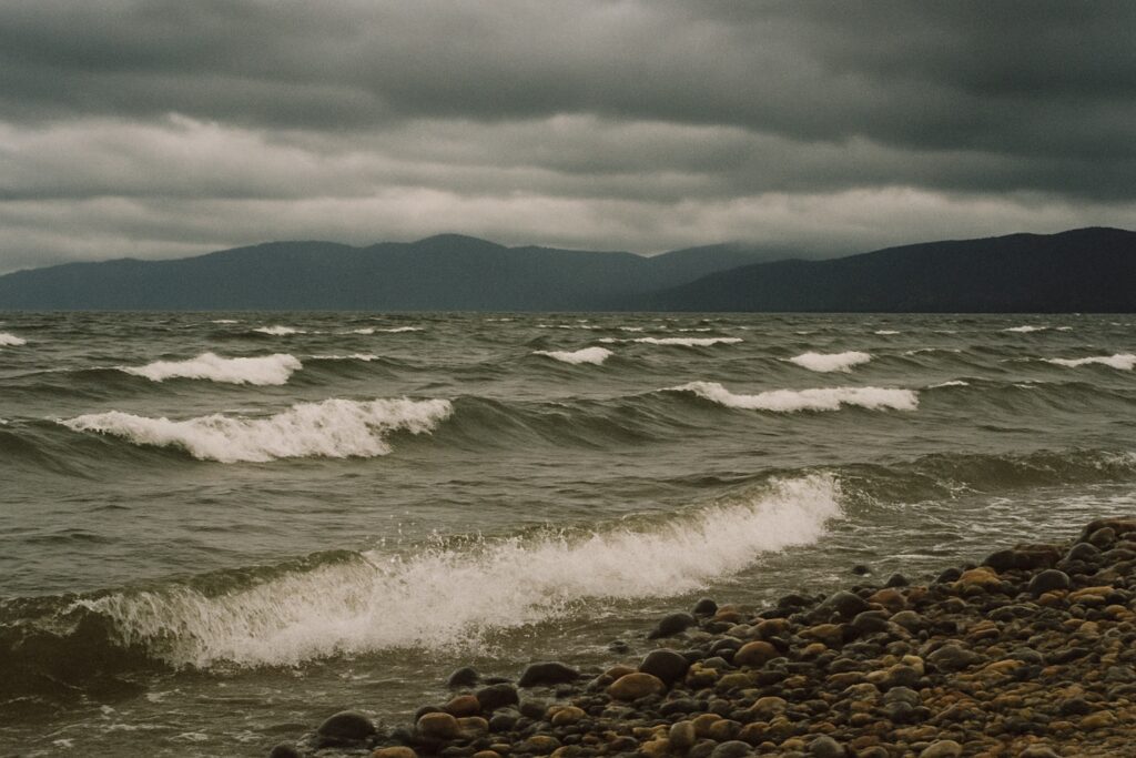 Graues, aufgewühltes Wasser mit vielen Schaumkronen in einer weiten Bucht, gesehen von einem flachen Kiesufer