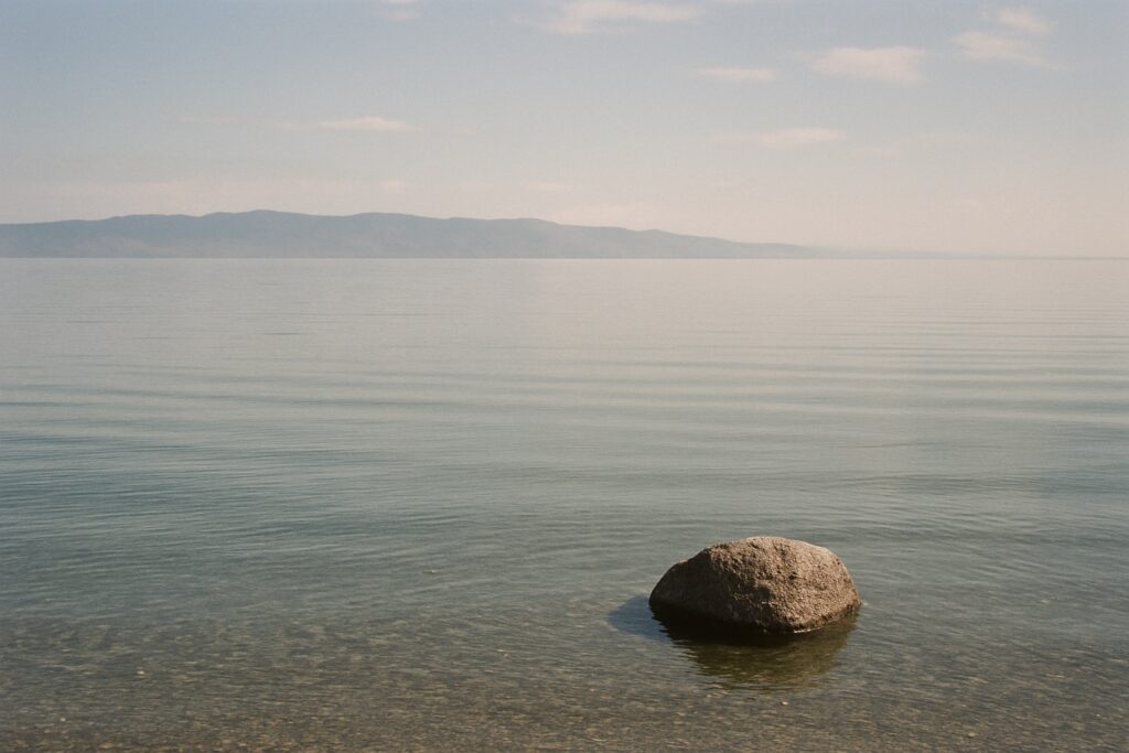 Weite, ruhige Wasserfläche bis zum Horizont mit einem fernen niedrigen Bergufer und einem Felsen im Vordergrund